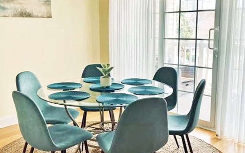 Round glass dining table with four teal upholstered chairs and place settings, bright windows with sheer curtains