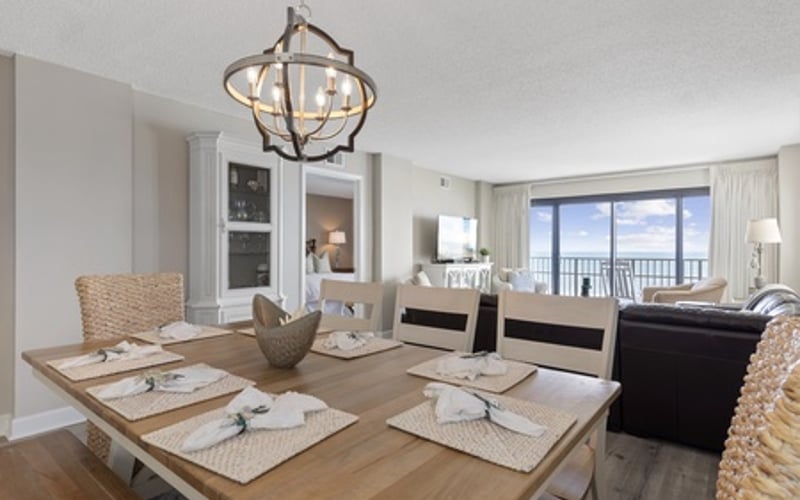 Open-plan dining and living room with wooden table, pendant light, and ocean view through large windows