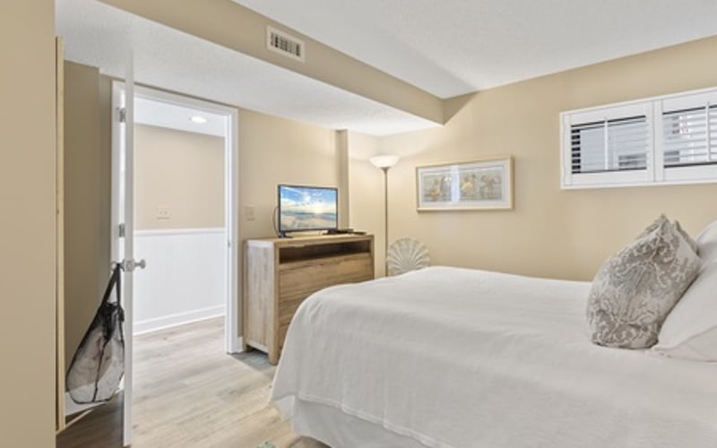 Bedroom with queen bed, wooden desk with monitor, cream walls, and glass door to hallway
